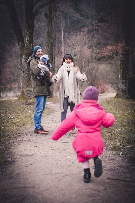 Mädchen laüft in die Arme seiner Mutter. Der Vater steht mit Baby auf dem Arm daneben.