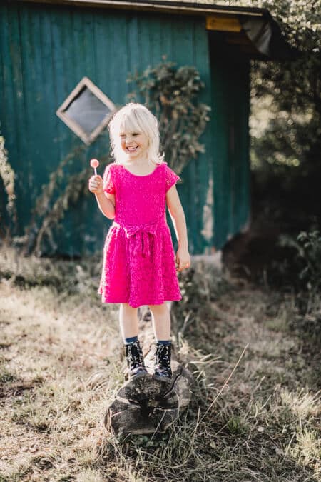 Mädchen mit pinkem Kleid und Lolli in der Hand auf einem Baumstamm