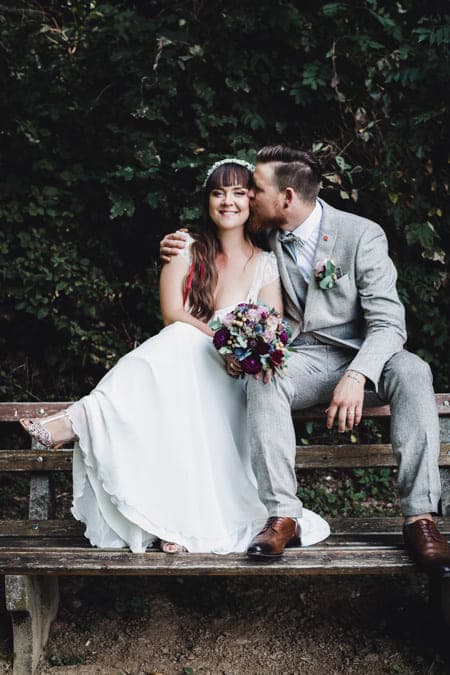 Brautpaar sitzt auf der Lehne einer Bank. Der Mann küsst seine Frau.