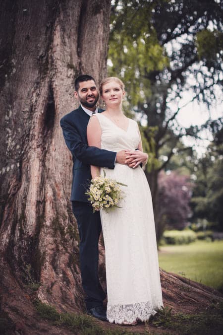 Brautpaar lehnt unter einem großen Baum am Baumstamm