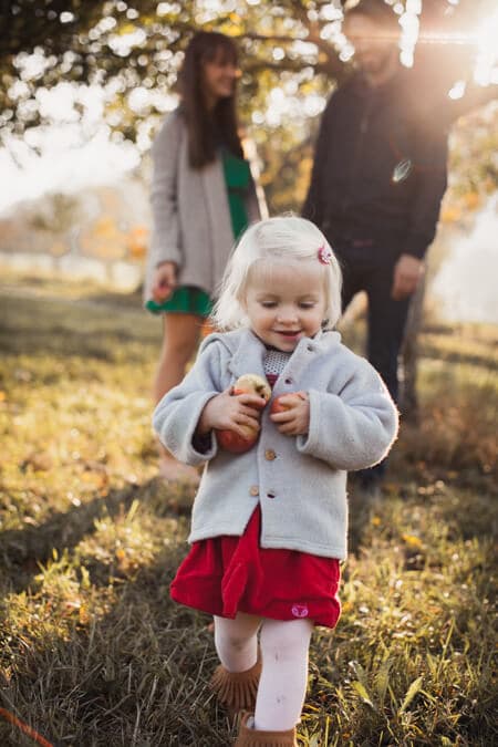 Mädchen mit zwei Äpfeln in den Händen. Im Hintergrund sind die Eltern.