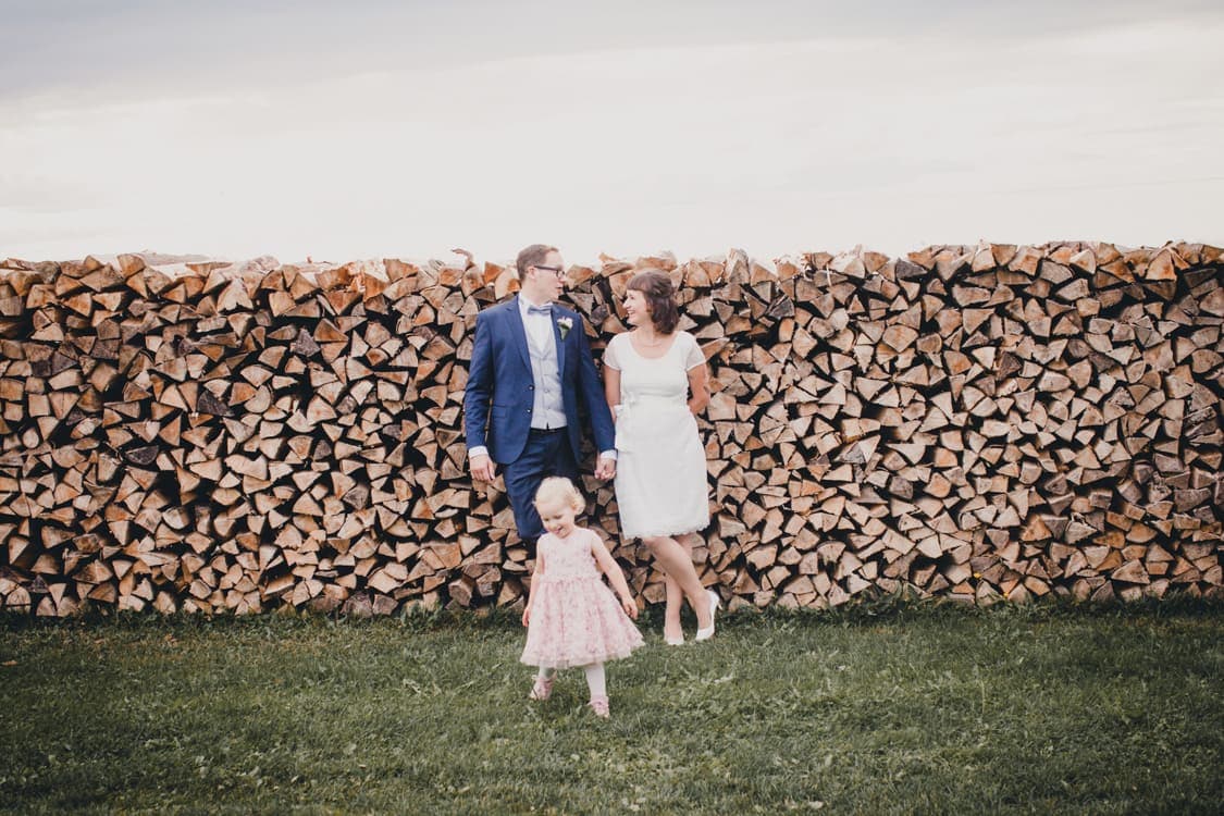 Brautpaar leht an Holzscheit und Tochter läuft im Vordergrund
