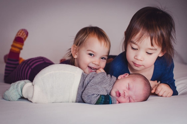 Baby liegt auf bäuchlings auf dem Bett. Seine Geschwister liegen dahinter und betrachten es.