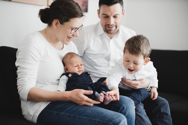 Familie sitzt auf dem Sofa. Junge greift nach den Füßen vom Baby.