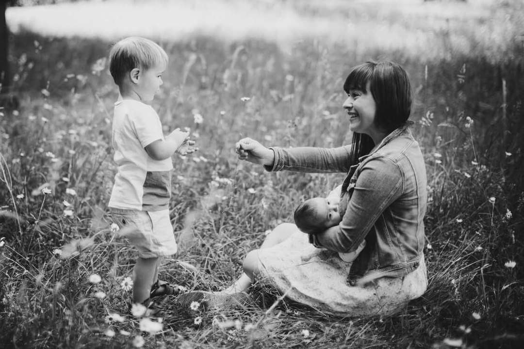 Mutter sitzt mit Baby auf einer Wiese. Der große Sohn pflückt der Mutter eine Blume.