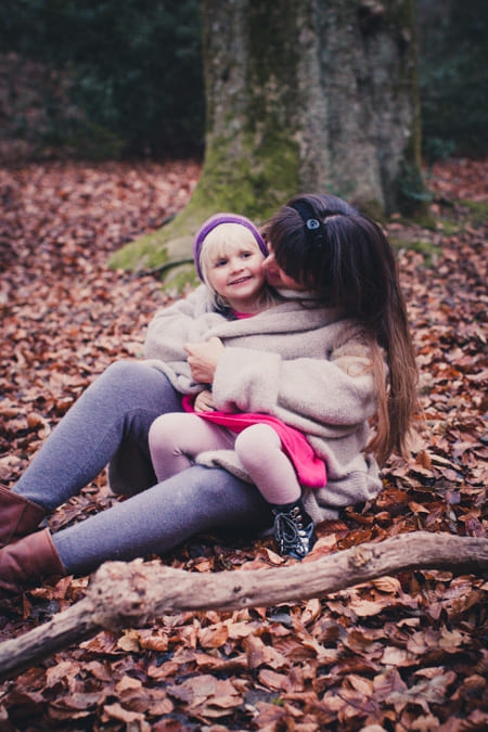 Mama sitzt mit ihrer Tochter auf dem Boden voller Blätter und nimmt sie unter ihren Strickmantel