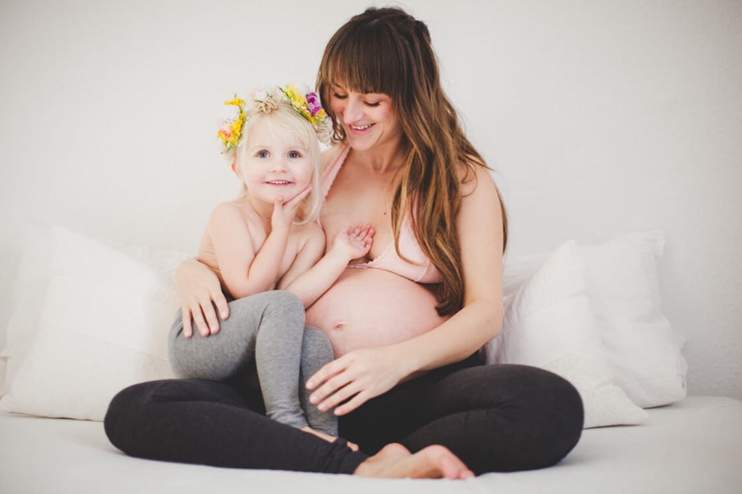 Tochter mit Blumenkranz sitzt bei Mama mit Babybauch auf dem Schoß