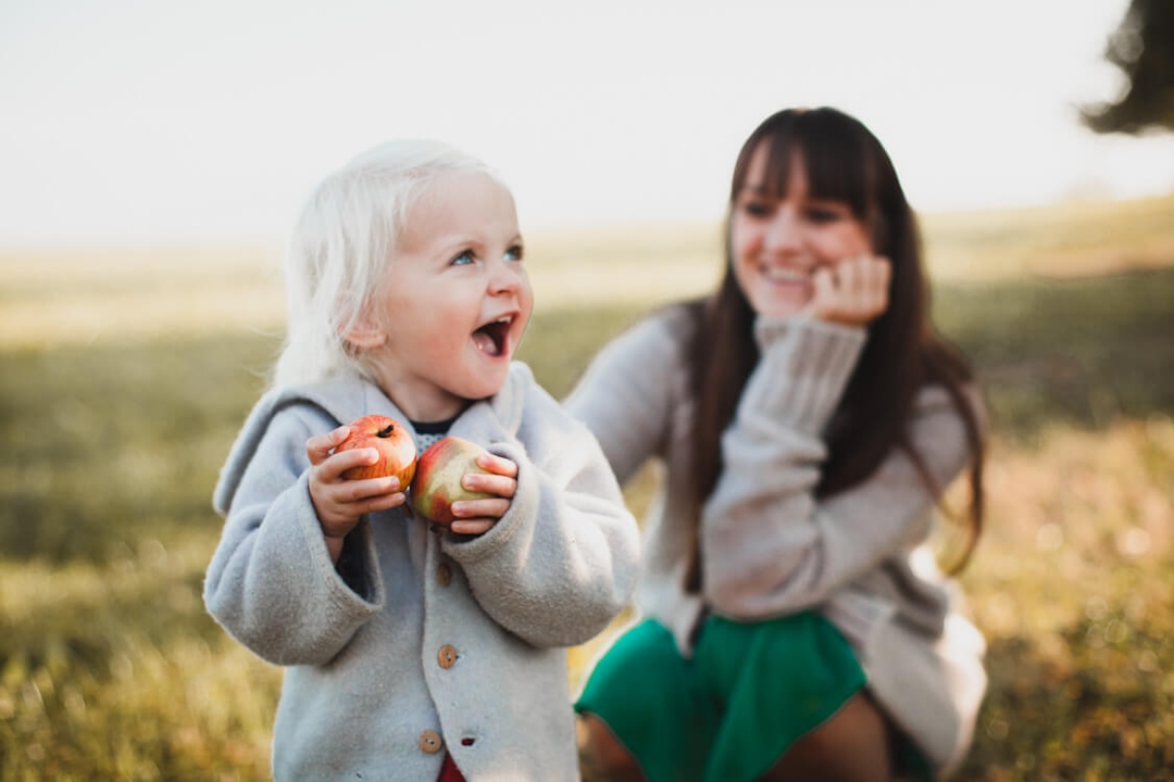 Kind mit zwei Äpfeln in den Händen und Mutter im Hintergrund