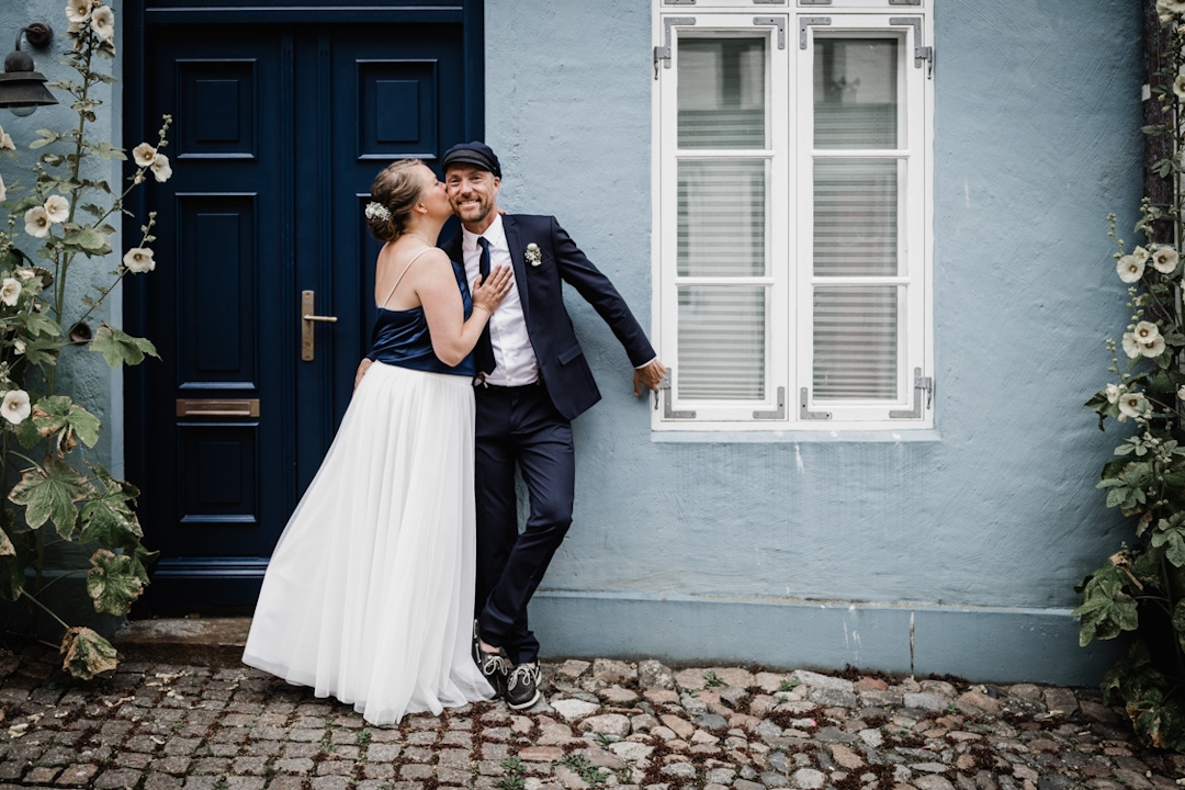Braut kuesst Braeutigam auf die Wange vor einem blauen Haus